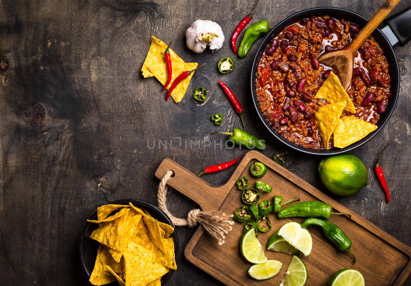 Chili con carne in frying pan on dark wooden background. Ingredients for making Chili con carne. Space for text. Top view. Chili with meat, nachos, lime, hot pepper. Mexican/Texas traditional dish