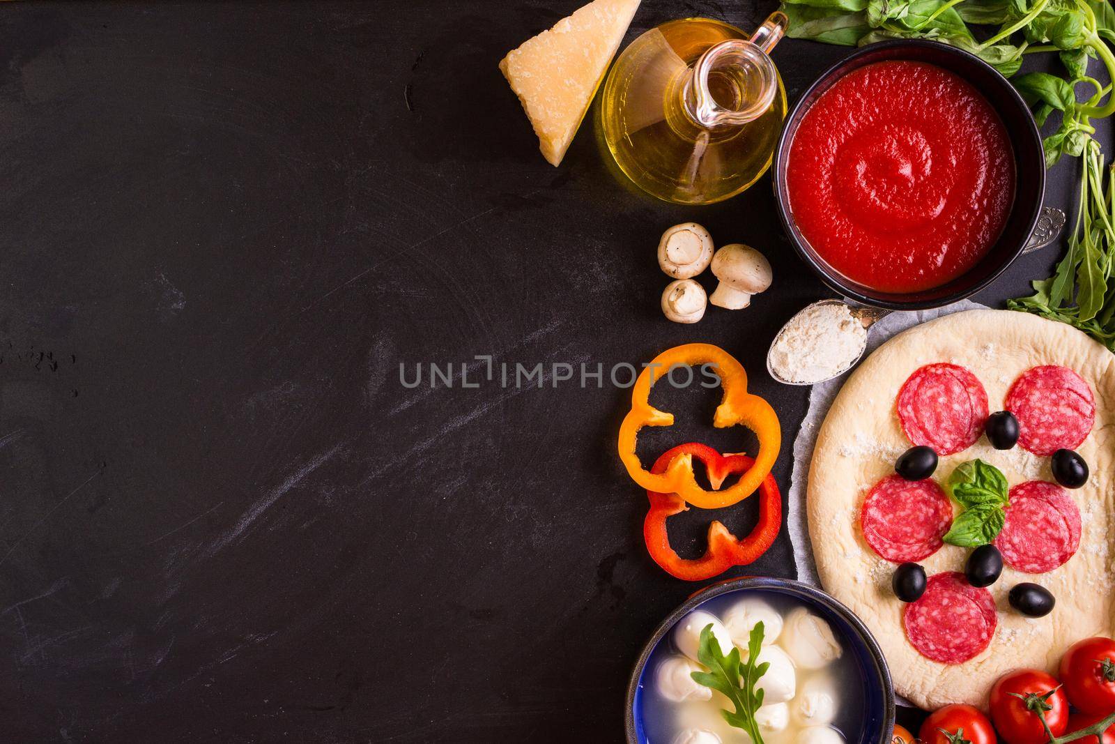 Pizza making background. Ingredients for making pizza. Space for text. Pizza dough, flour, cheese, mozzarella, tomatoes, basil, pepperoni, olives and rolling pin over black chalkboard. Top view