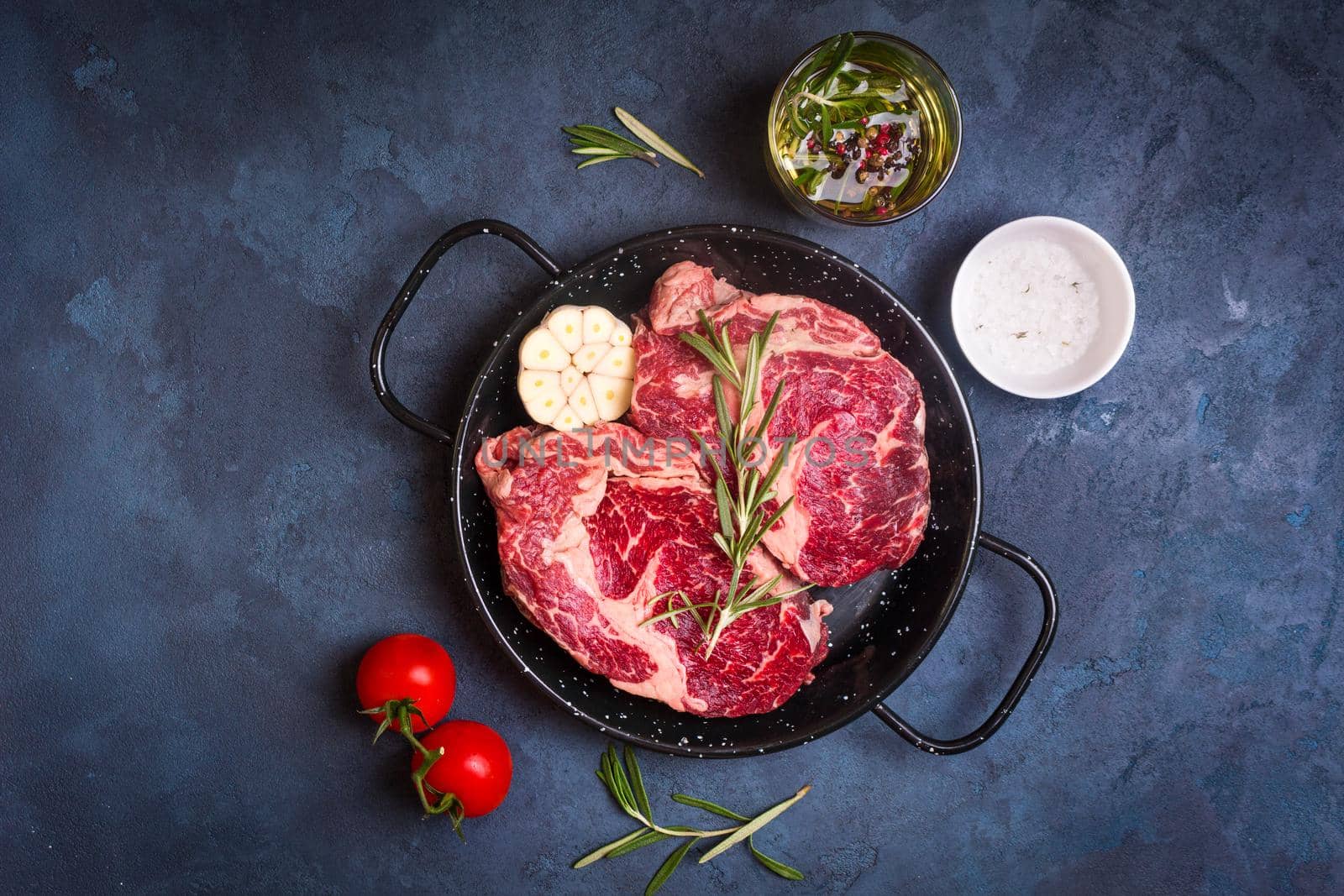 Raw juicy steaks with seasonings in a black pan ready for roasting on rustic concrete background. Fresh marbled meat steaks with herbs, garlic, olive oil, pepper, salt and tomatoes. Top view