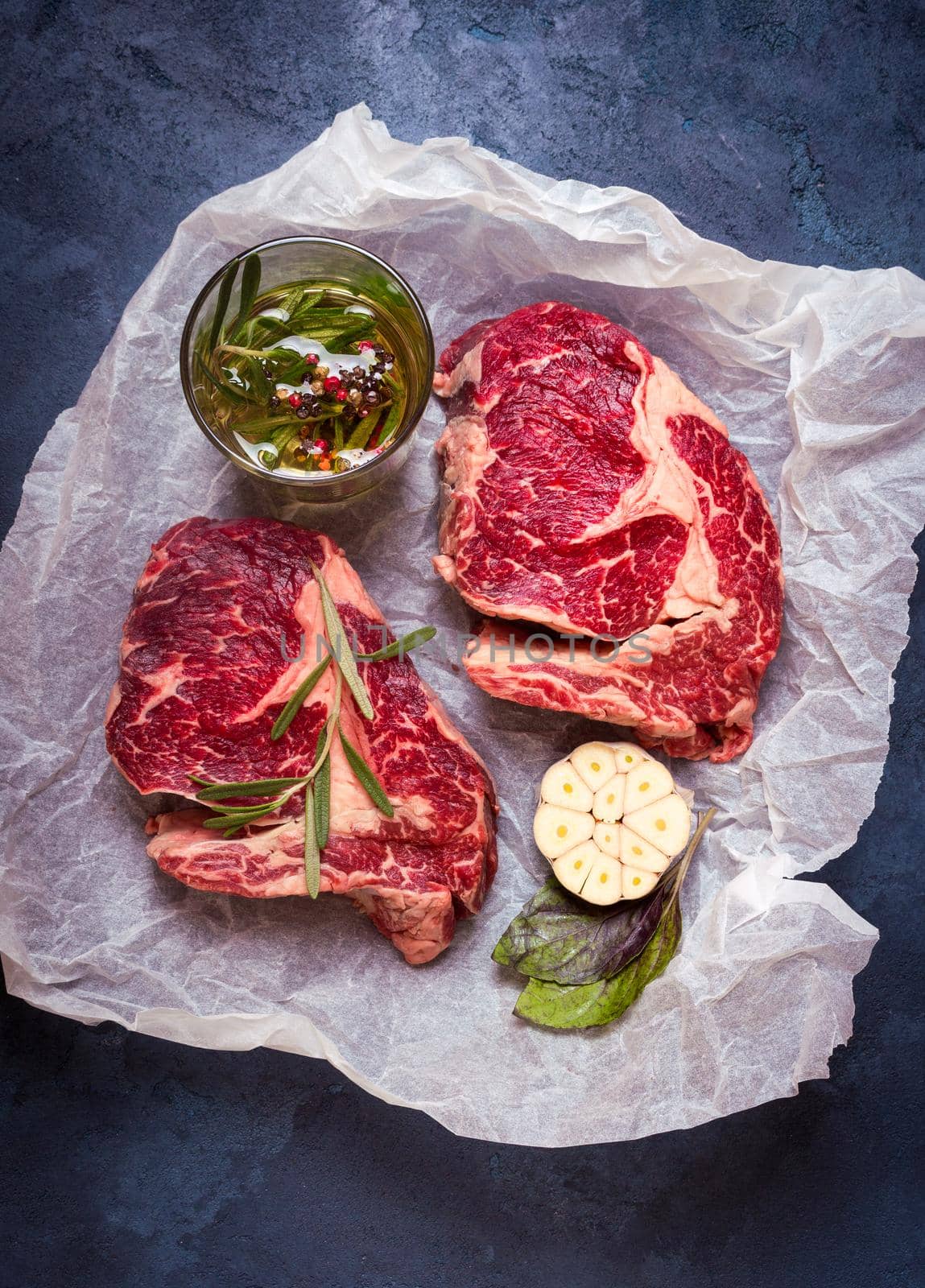 Raw juicy steaks with seasonings on a baking paper ready for roasting on rustic concrete background. Fresh marbled meat steaks with herbs, garlic, olive oil, colorful whole pepper, rock salt. Top view