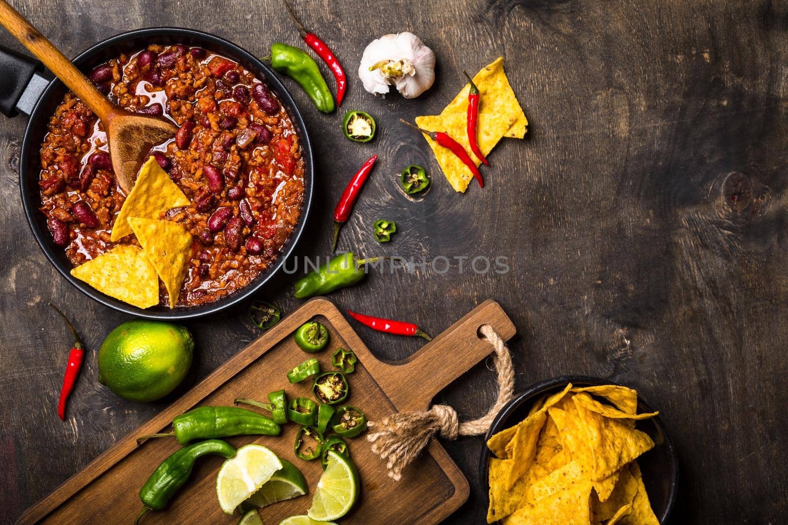 Chili con carne in frying pan on dark wooden background. Ingredients for making Chili con carne. Space for text. Top view. Chili with meat, nachos, lime, hot pepper. Mexican/Texas traditional dish