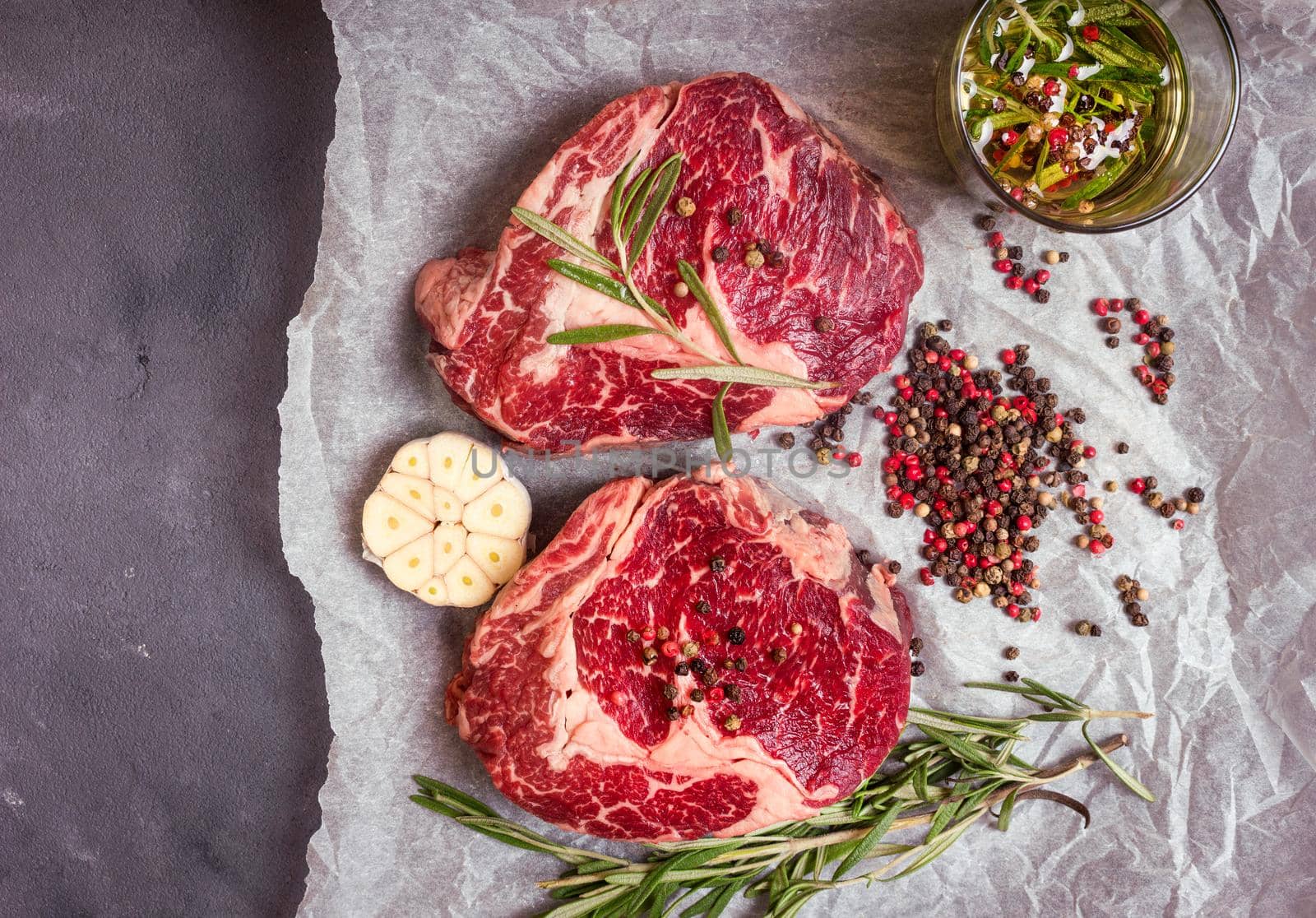 Raw juicy steaks with seasonings on a baking paper ready for roasting on rustic concrete background. Fresh marbled meat steaks with herbs, garlic, olive oil, colorful whole pepper, rock salt. Top view