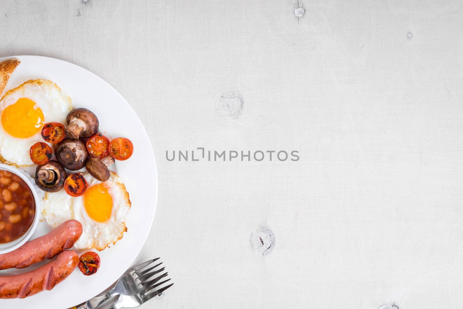 Full english breakfast with fried eggs, tomatoes, sausages, bacon, mushrooms, toasts and beans. Breakfast on a white plate with forks on the white wooden table. Space for text. Background