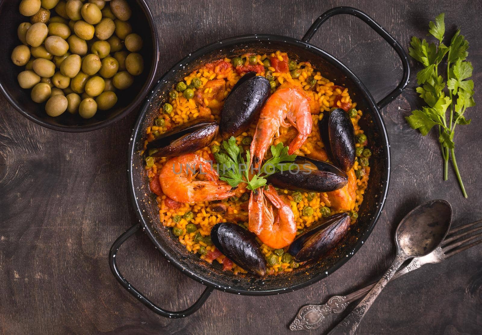 Paella in black pan with rice, shrimps, mussels, squid and meat, bowl with olives and vintage cutlery. Seafood paella, traditional spanish dish. Paella on rustic table. Selective focus. Top view