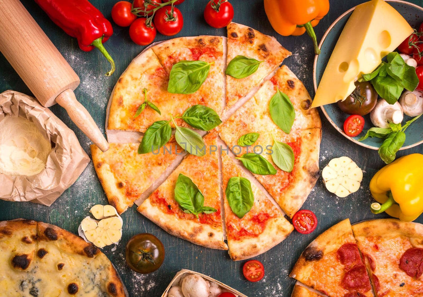 Delicious sliced pizza and ingredients for making pizza. Flour, cheese, tomatoes, basil, pepperoni, mushrooms and rolling pin over wooden background. Top view. Pizza ready to eat