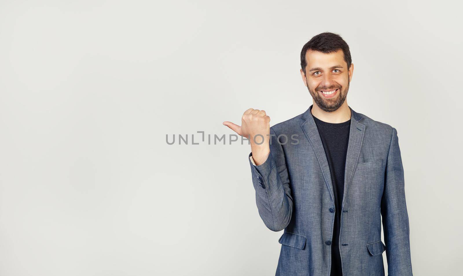 A young man with a beard smiling with a happy face, looking and pointing to the side with his thumb at a place for advertising, look here, on isolated background.