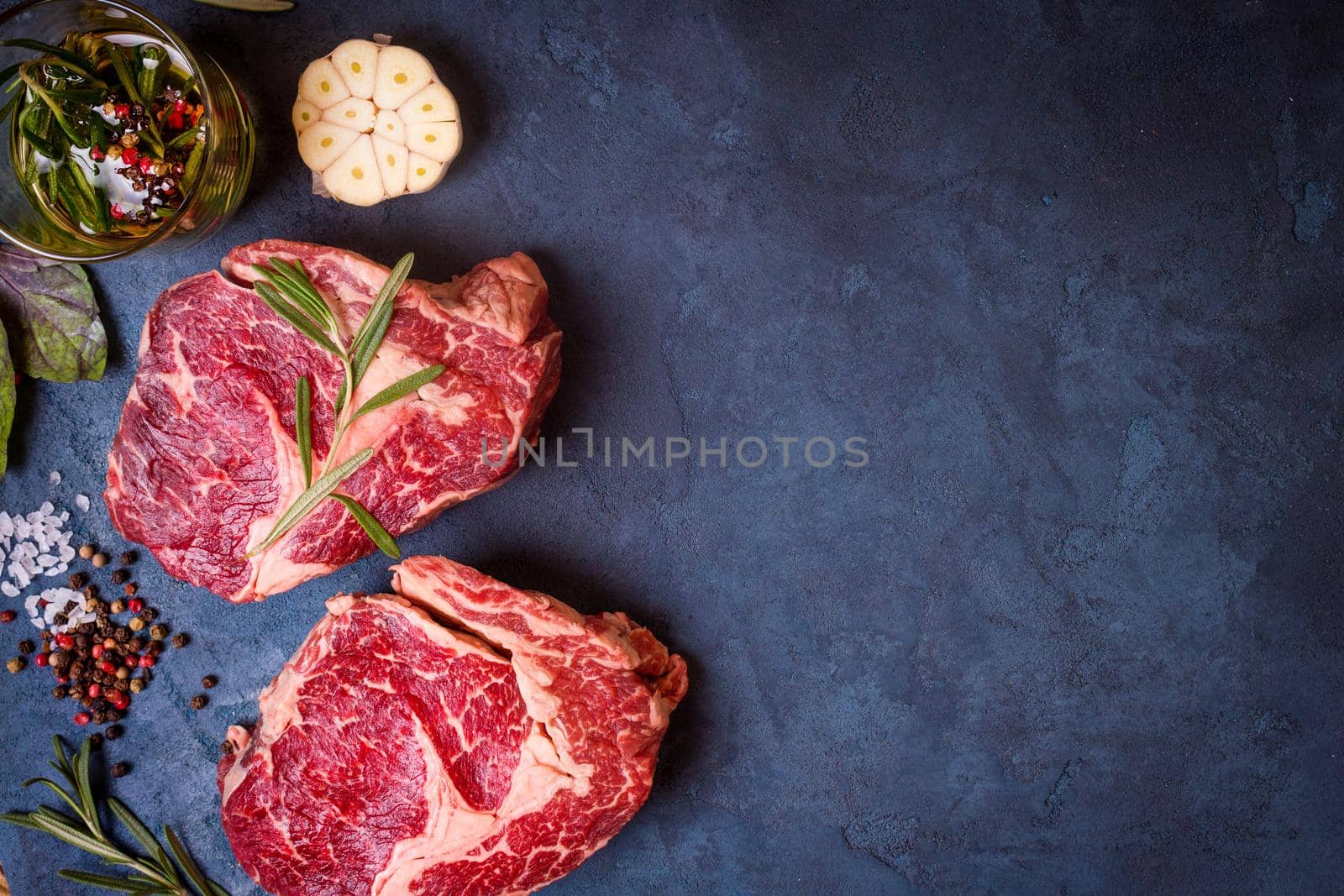 Raw juicy ribeye steaks with seasonings ready for roasting on rustic concrete background. Fresh marbled meat steaks with herbs, garlic, olive oil, whole pepper and rock salt. Space for text. Top view