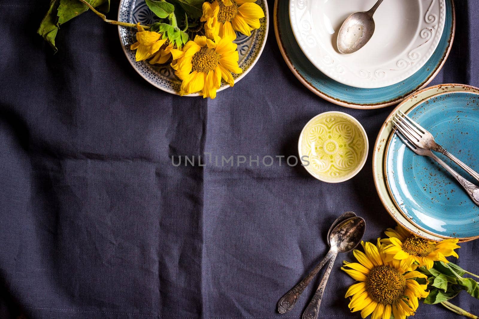 Vintage multicolored empty plates and bowls on a dark gray linen tablecloth with sunflower. With antique spoons and forks. Space for text. Table setting. Shabby chic/retro style. Top view. Copy space