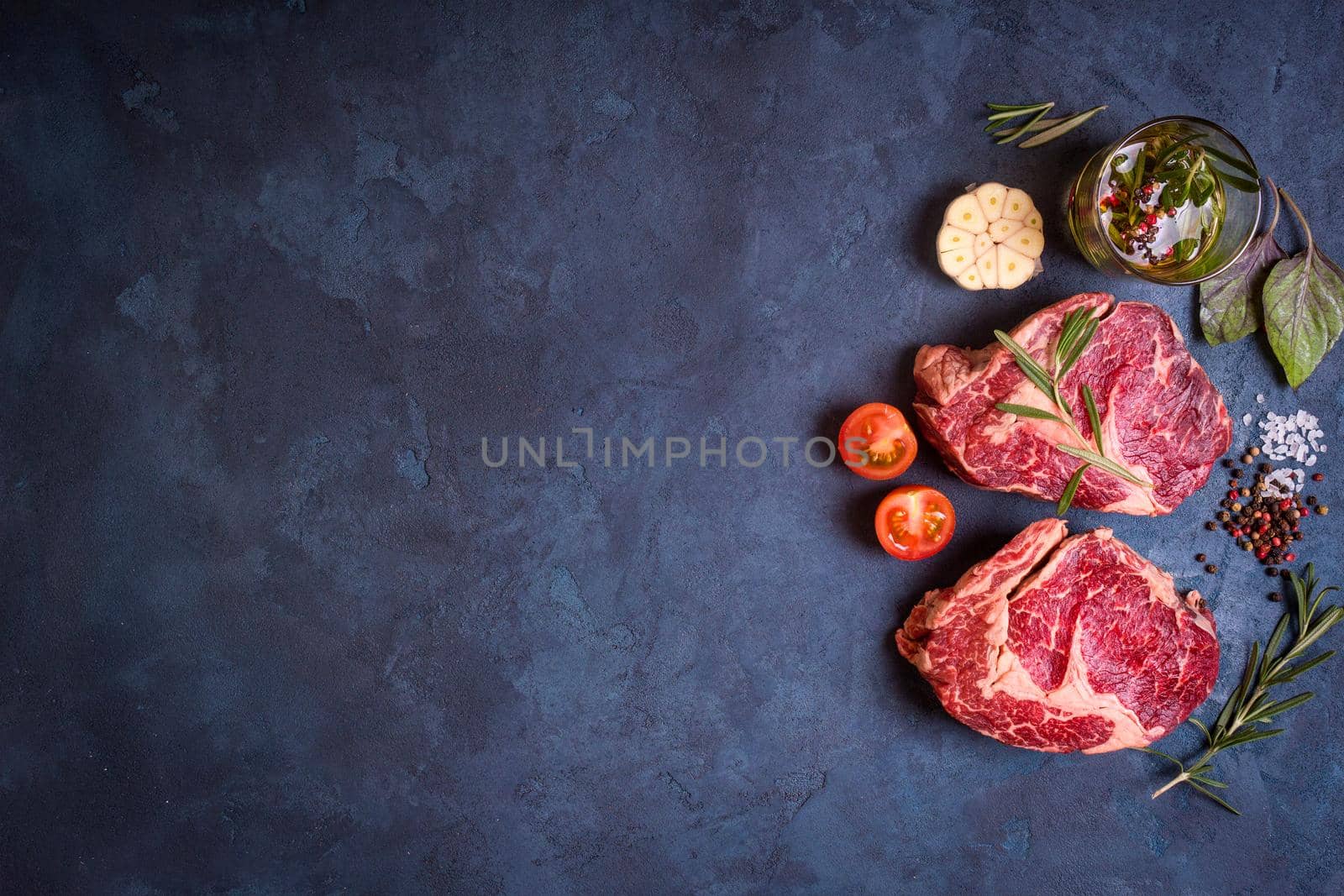 Raw juicy ribeye steaks with seasonings ready for roasting on rustic concrete background. Fresh marbled meat steaks with herbs, garlic, olive oil, pepper, salt and tomatoes. Space for text. Top view