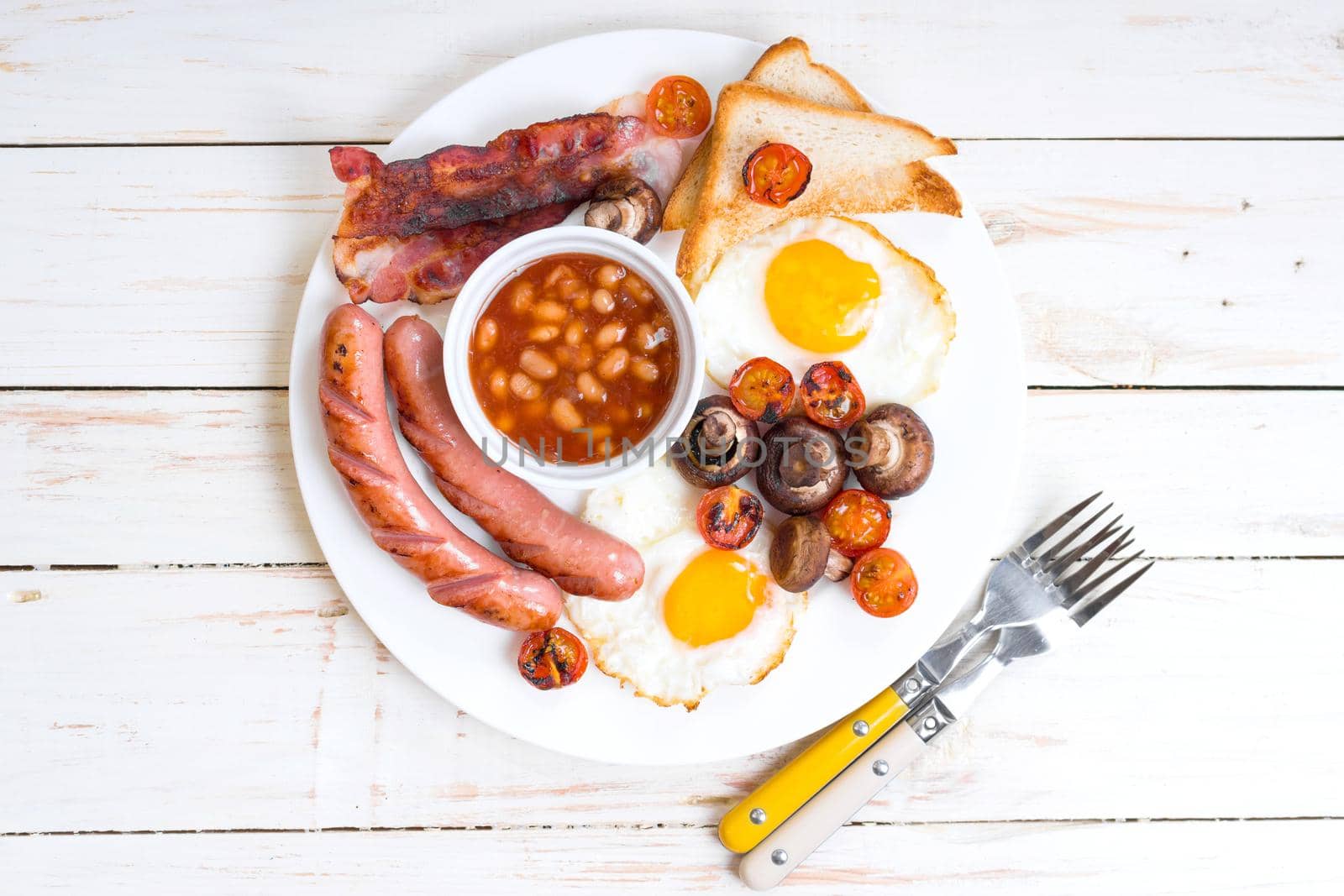 Full english breakfast with fried eggs, tomatoes, sausages, bacon, mushrooms, toasts and beans. Breakfast on a white plate with forks on the white wooden table. Top view