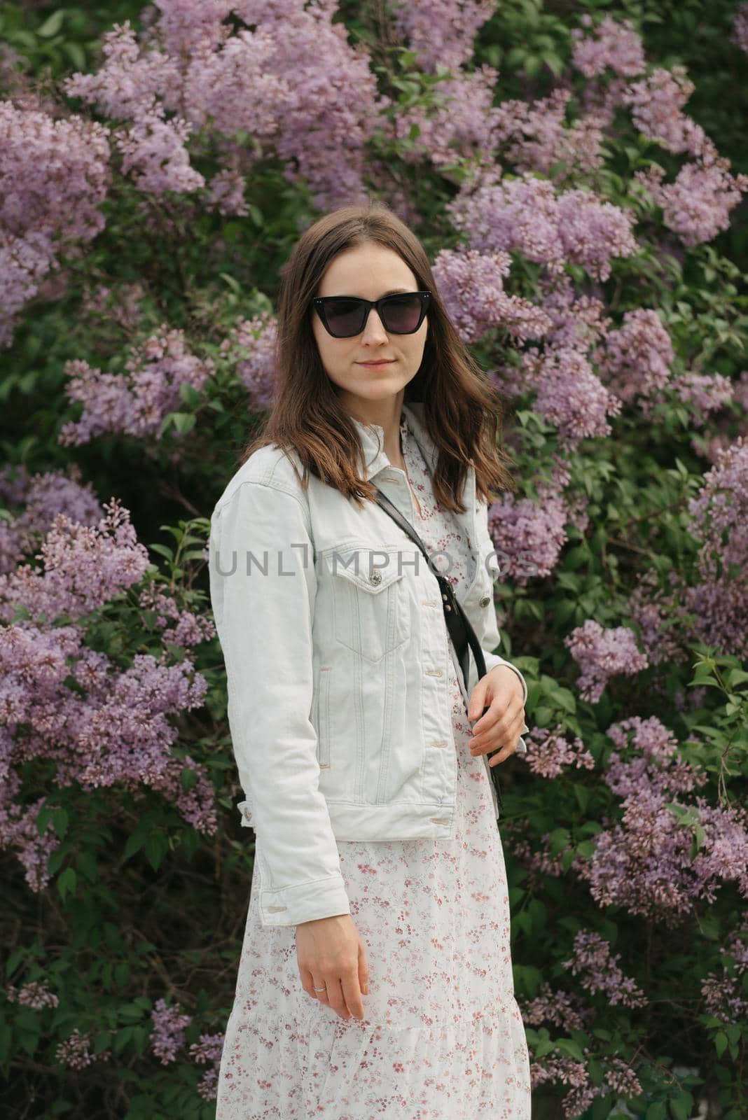 A woman is staying near the lilac bush in the village green. A lady in black sunglasses is posing in the woodland.