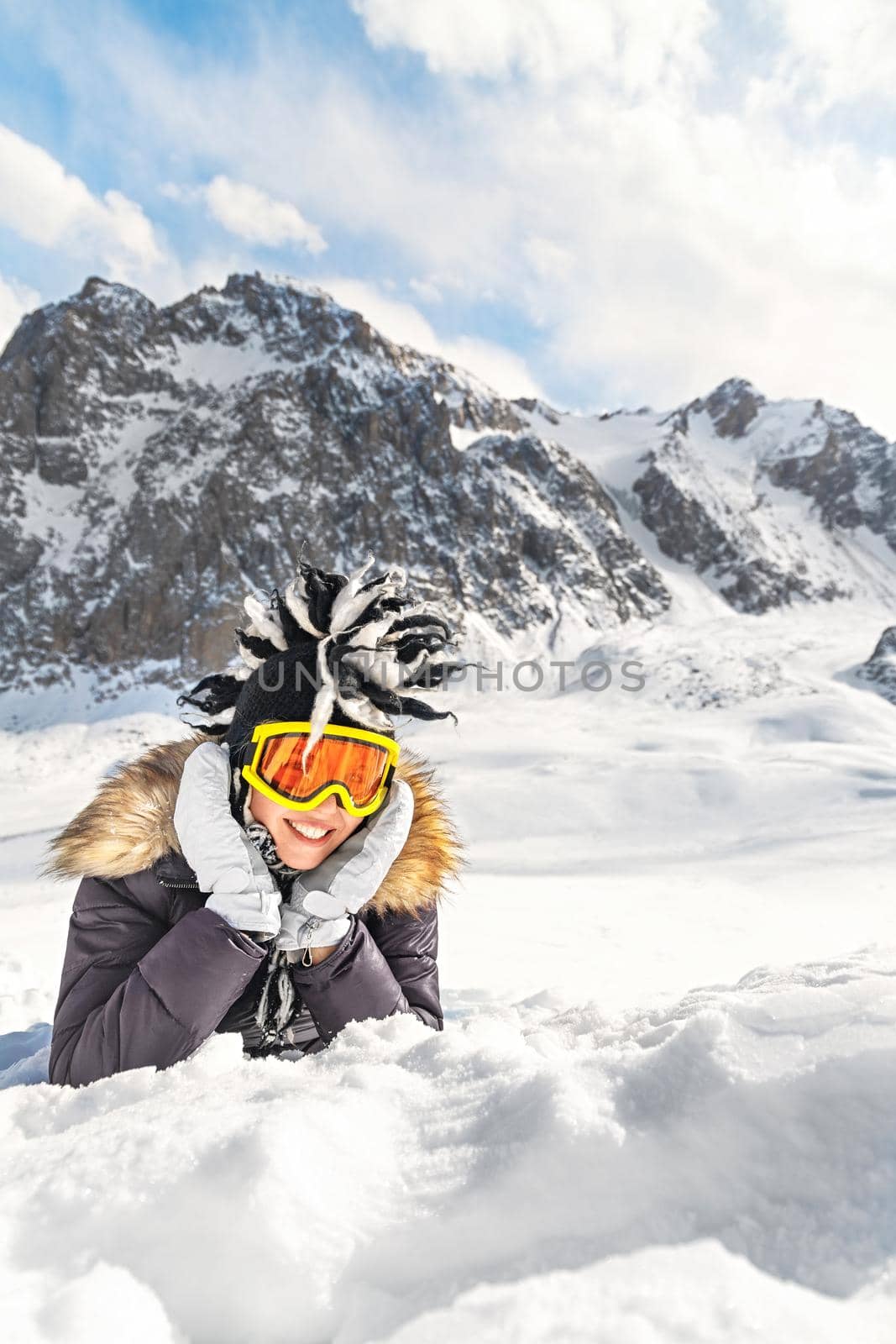 Cute young woman dressed in winter clothes portrait, glasses and winter funny hat, resting on snow top of the mountain. Winter travelling scene, wanderlust concept. Copy space, vertical