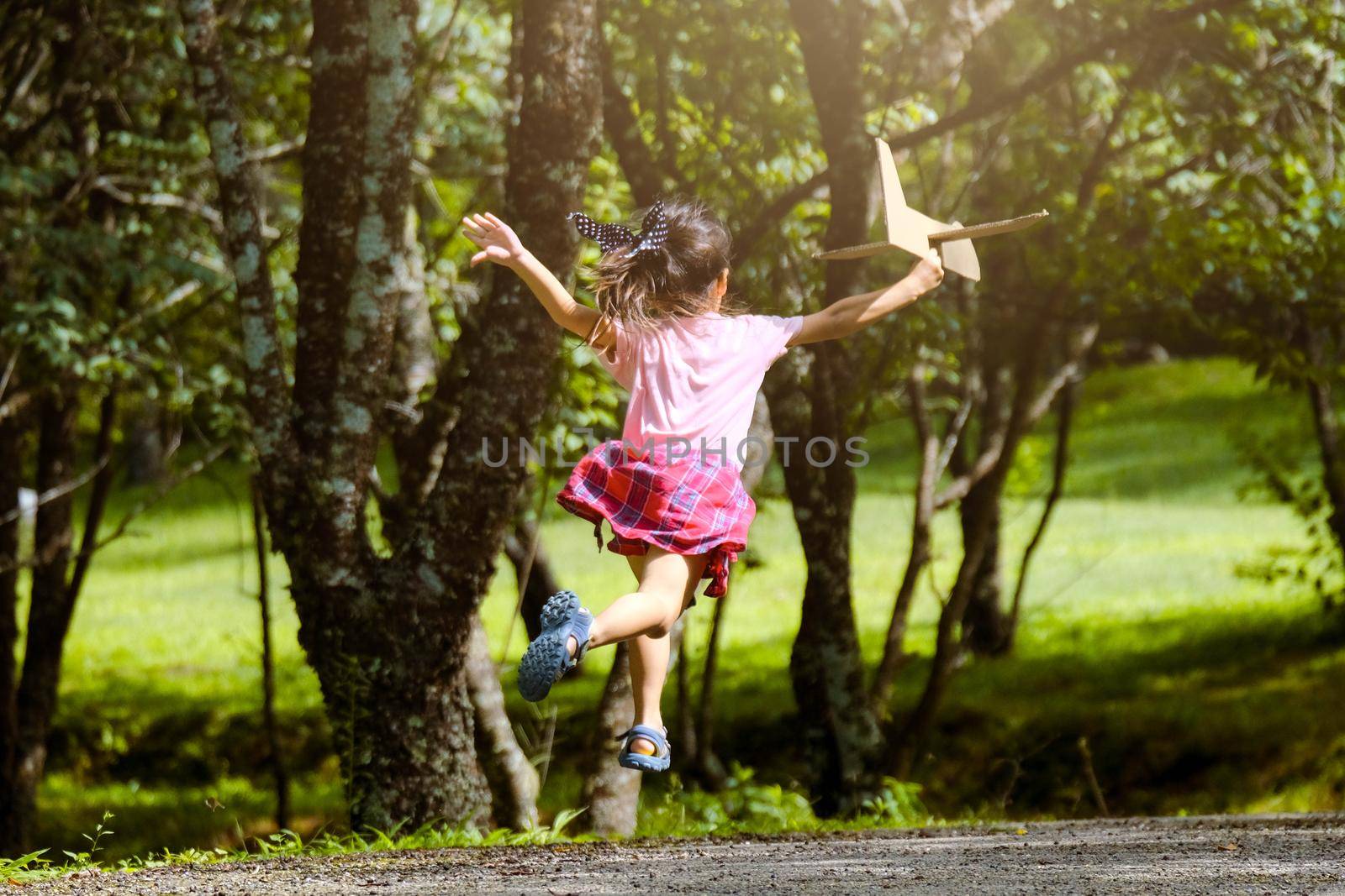 Happy little girl playing with airplanes made of cardboard in summer park. Cute girl with a toy airplane running on the road in the park. Happy childhood concept. by TEERASAK