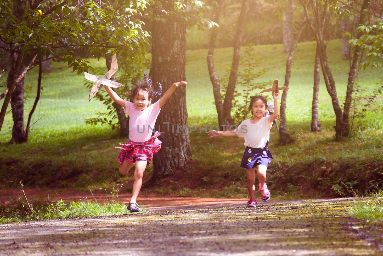 Happy children playing with cardboard planes in the summer garden. Two little sisters with toy planes running on the road in the park. happy childhood concept. by TEERASAK