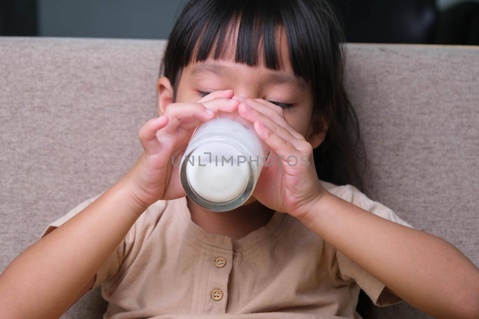 Portrait of a cute Asian little girl holding a glass of milk sitting on the sofa at home. Small girl at home with smiling face, feeling happy enjoying drinking milk and looking at camera. by TEERASAK
