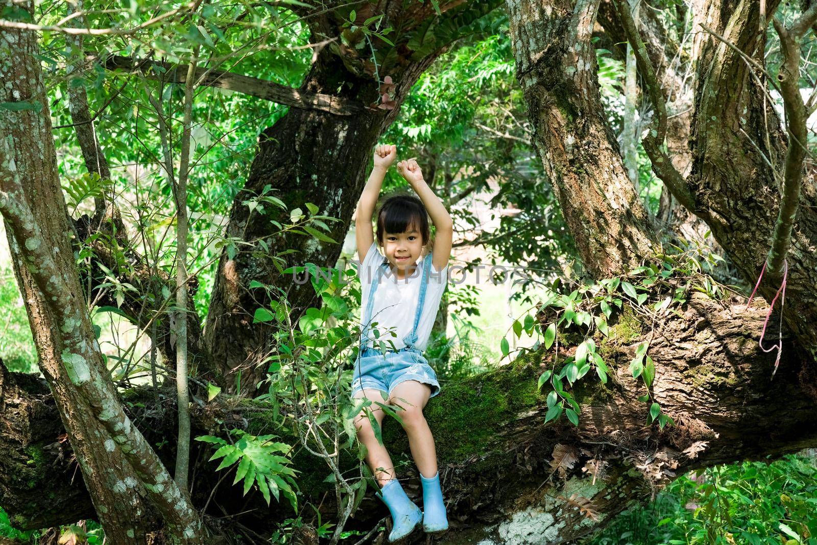 Cute little girl in boots playing in the garden, climbing a tree. Happy girl relaxing on tree branch. by TEERASAK