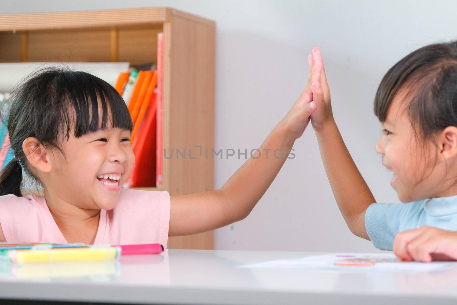 Little girl giving a hi-five with to her friends sitting in the classroom. Cute little sisters taking lessons for home schooling. Elementary school children enjoy learning together. Back to school