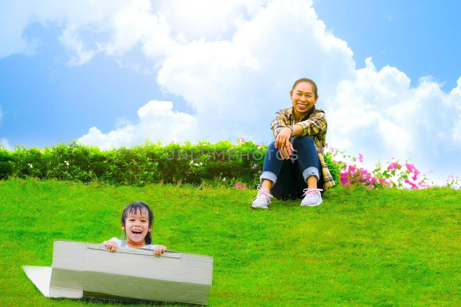 Smiling little girl sat on a cardboard box sliding down a hill at the Botanical Garden and her mother smiled as she looked at her. Happy childhood concept.
