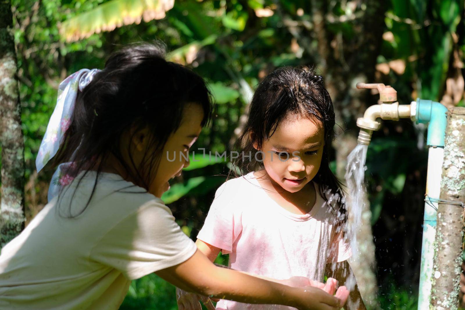 Two little sisters washing their hands at the outdoor faucet. Two cute girls turn on the faucet to wash their hands in the park. by TEERASAK