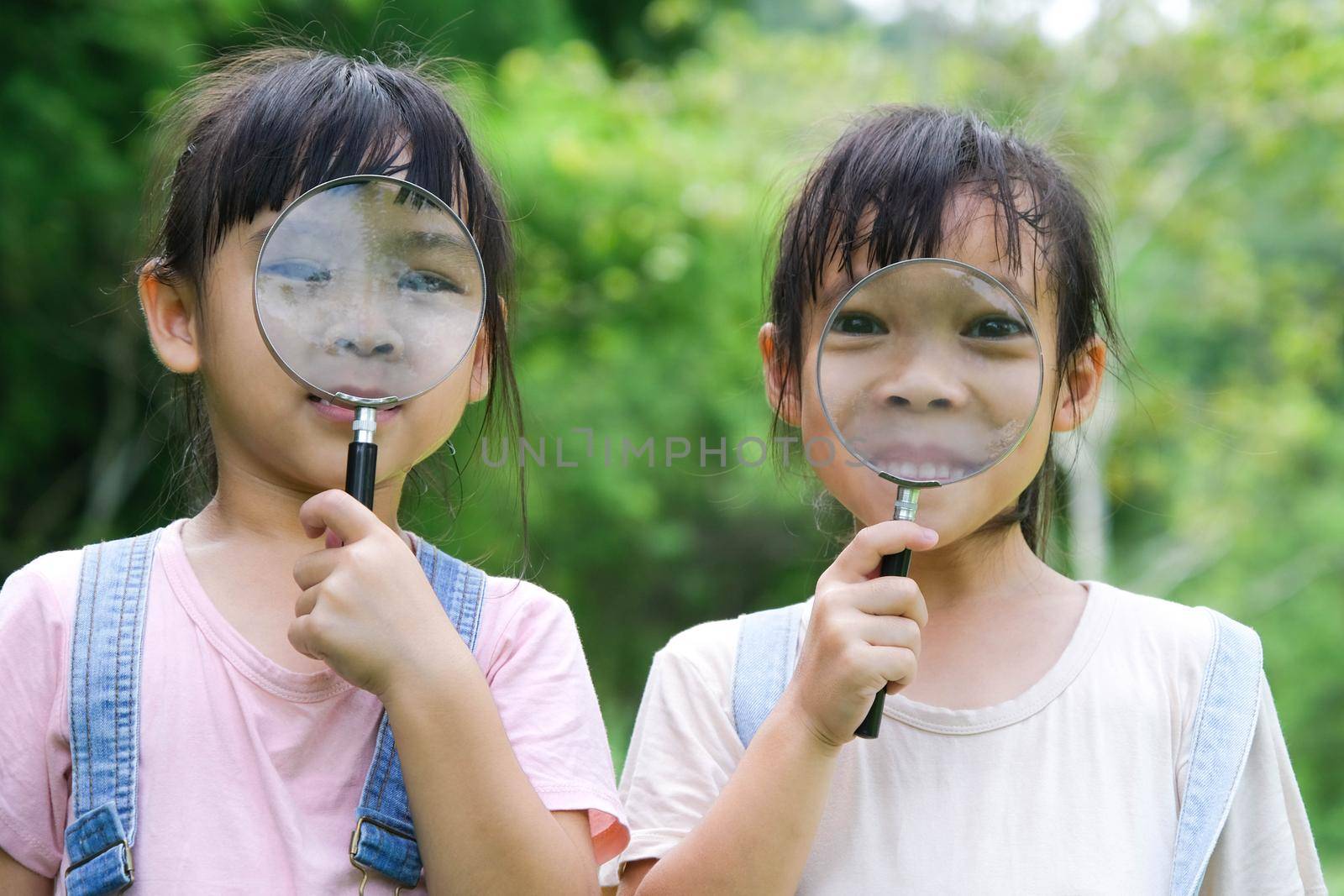 Children learn and explore nature with an outdoor magnifying glass. Curious child looks through a magnifying glass at the trees in the park.  Two little sisters playing with magnifying glass. by TEERASAK