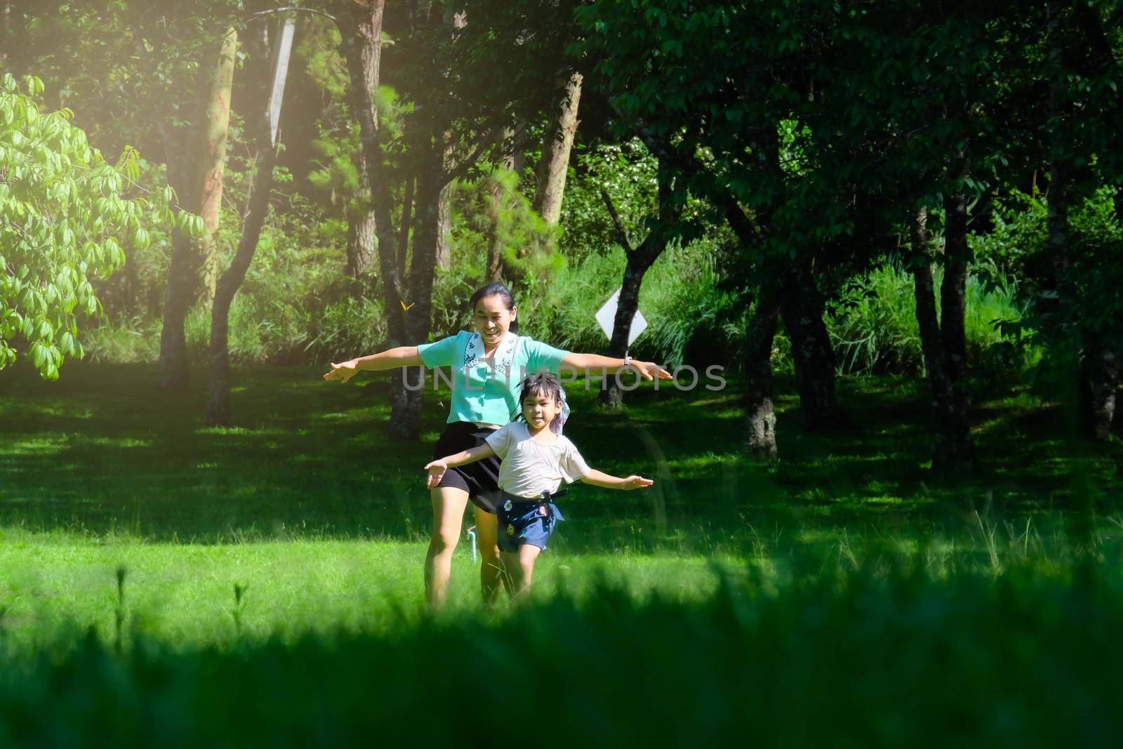 Cute little girl runs with her mother spreading her arms as if flying in a spring garden. Young mother playing with her daughter in the park. Happy family having fun in the park. by TEERASAK