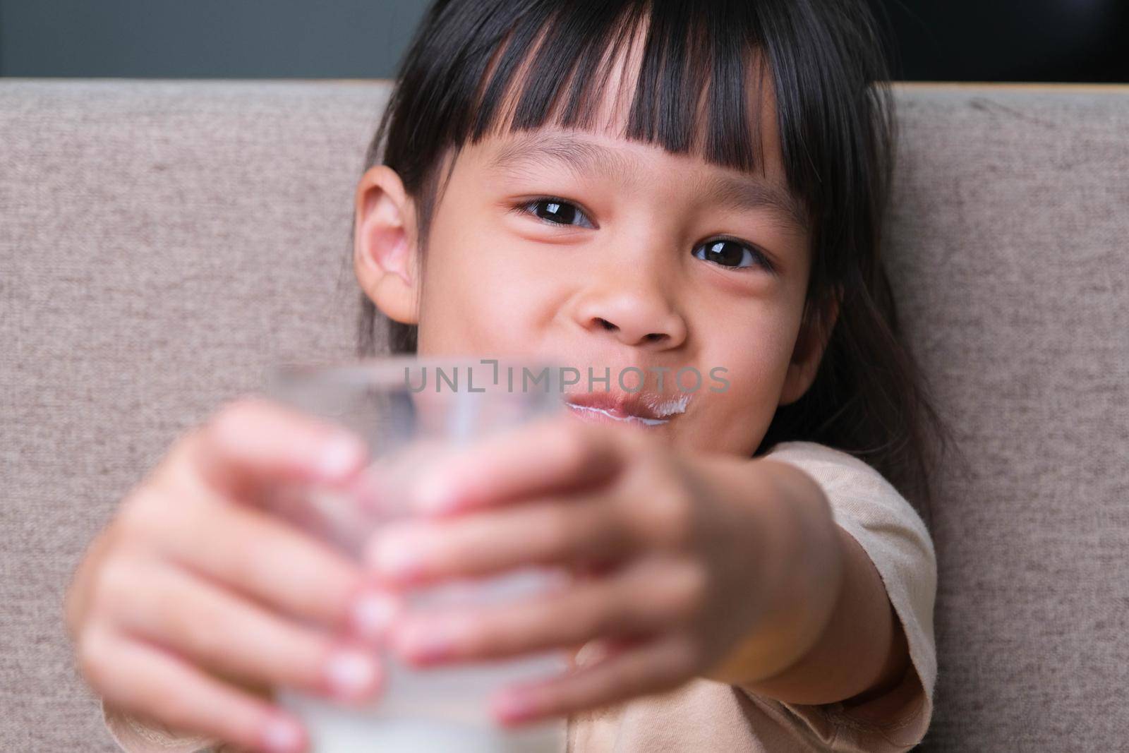 Portrait of a cute Asian little girl holding a glass of milk sitting on the sofa at home. Small girl at home with smiling face, feeling happy enjoying drinking milk and looking at camera.