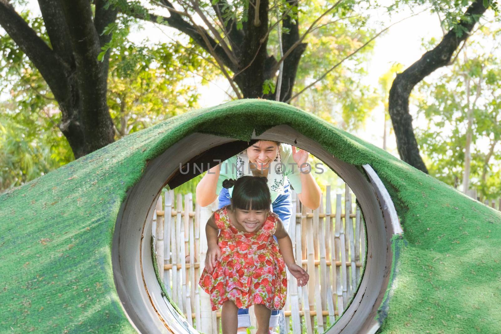 Cute little girl and young mother playing together in the mountains with tunnel in the outdoor playground. Healthy summer activity for children. by TEERASAK