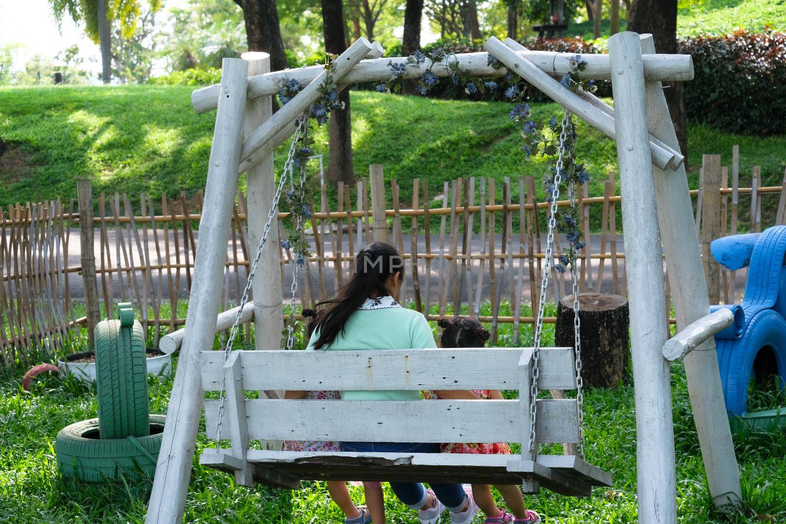 Rear view of little girls sitting on a swing under tree with mother in the summer park. Happy family having fun in the park.