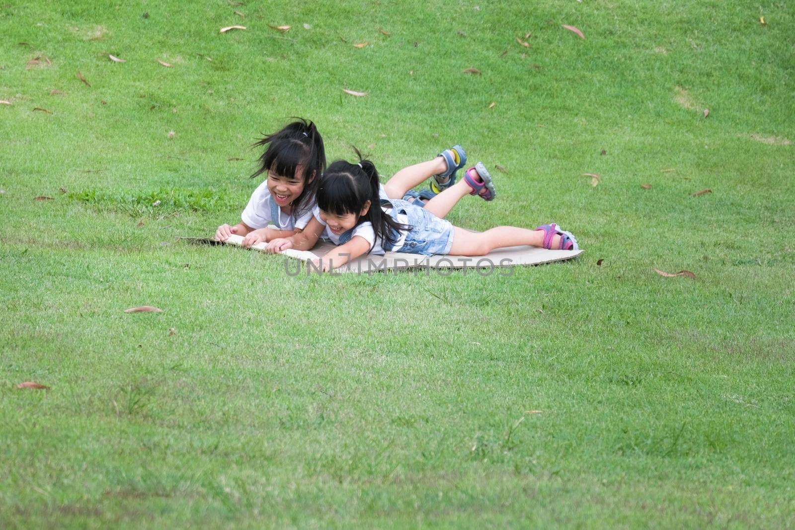 Two smiling little sisters lie prone on cardboard boxes sliding down the hill at the Botanical Gardens. The famous outdoor learning center of Mae Moh Mine Park, Lampang, Thailand. Happy childhood conc by TEERASAK