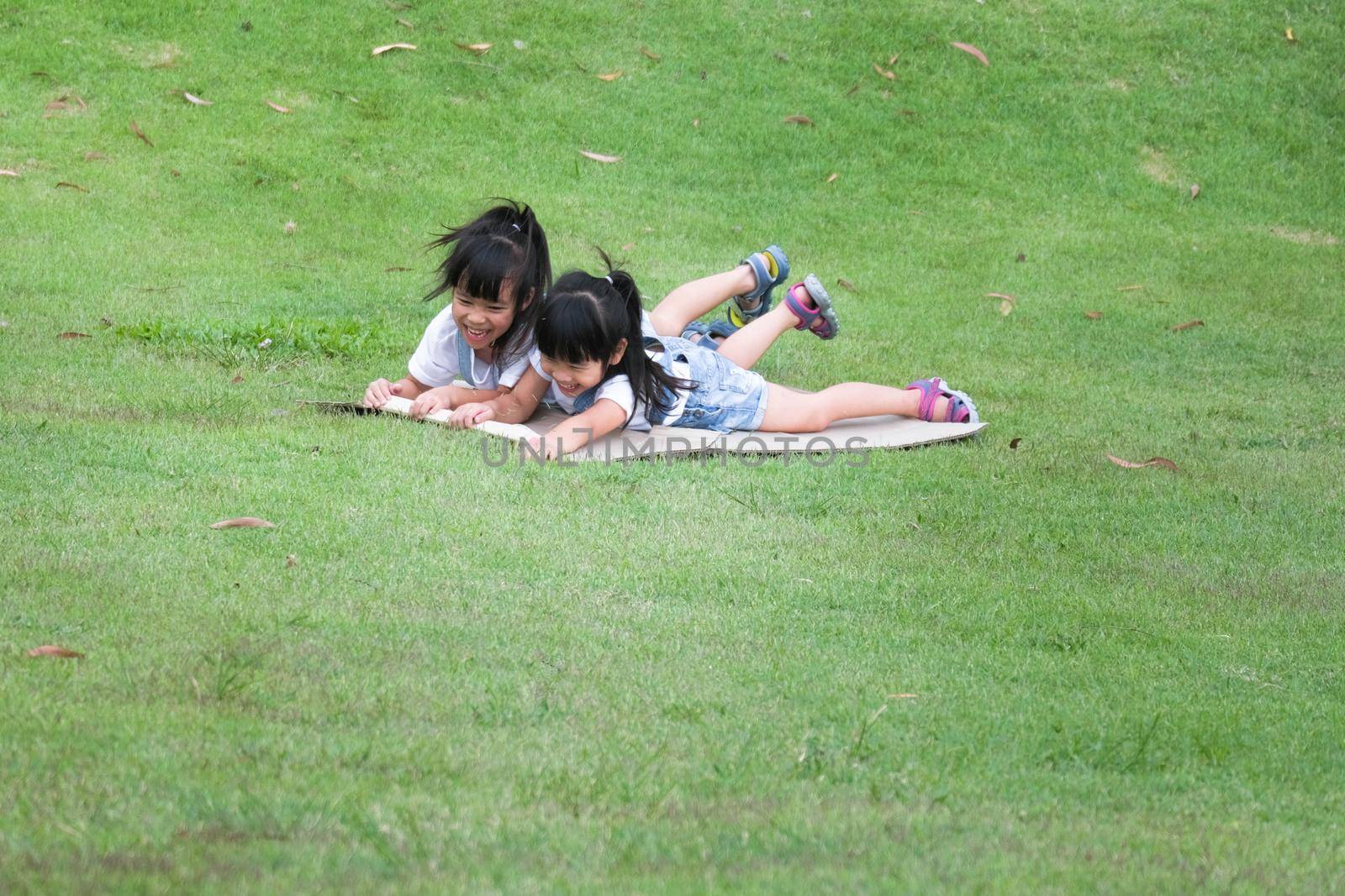 Two smiling little sisters lie prone on cardboard boxes sliding down the hill at the Botanical Gardens. The famous outdoor learning center of Mae Moh Mine Park, Lampang, Thailand. Happy childhood conc