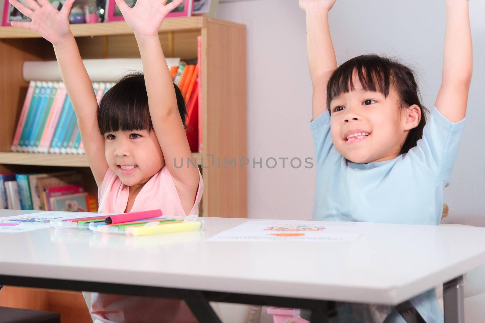 Happy kids with arms up sit in classroom. Cute girl taking lessons for home schooling Elementary school children enjoy learning together. Back to school concept. by TEERASAK