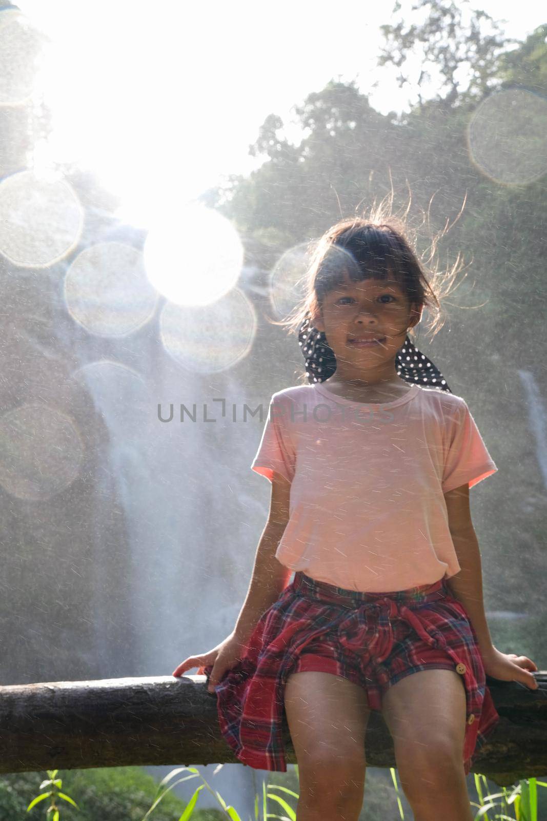 Cute Asian girl sits on a log near Wachirathan Waterfall, looks at camera and smiles in Inthanon National Park, Chiang Mai, Thailand. by TEERASAK