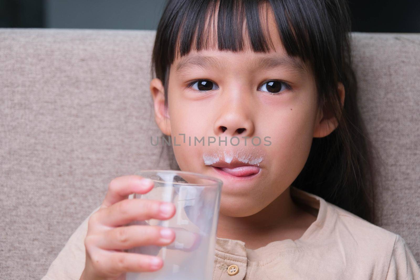 Portrait of a cute Asian little girl holding a glass of milk sitting on the sofa at home. Small girl at home with smiling face, feeling happy enjoying drinking milk and looking at camera.