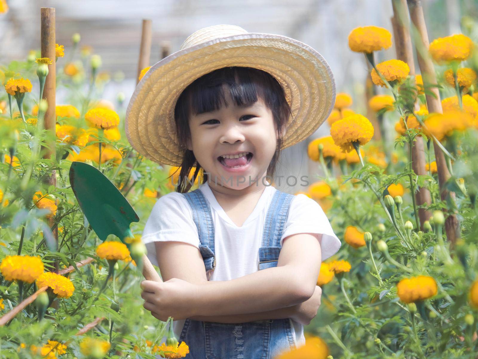 Cute little girl in hat holding garden tool shovel for planting flowers in the garden. A child helps mom in the garden, a little gardener. by TEERASAK
