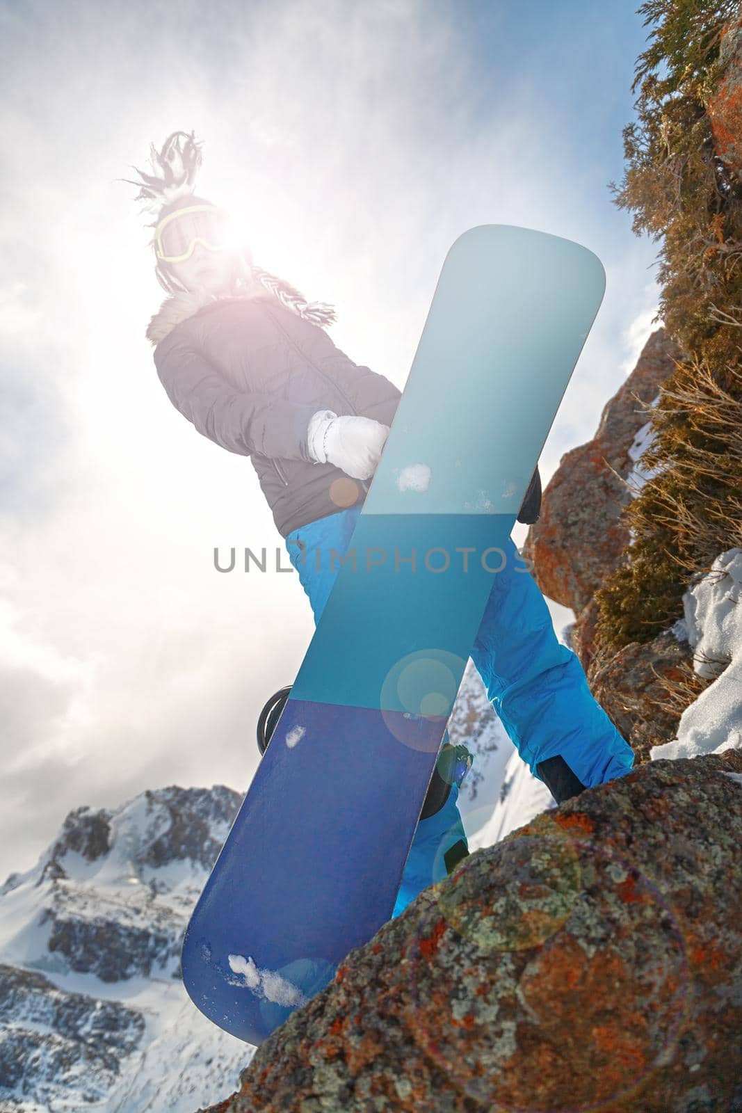 Beautiful young female snowboarder with blue snowboard and funny hat on the mountains. Look at camera. lens flare effect