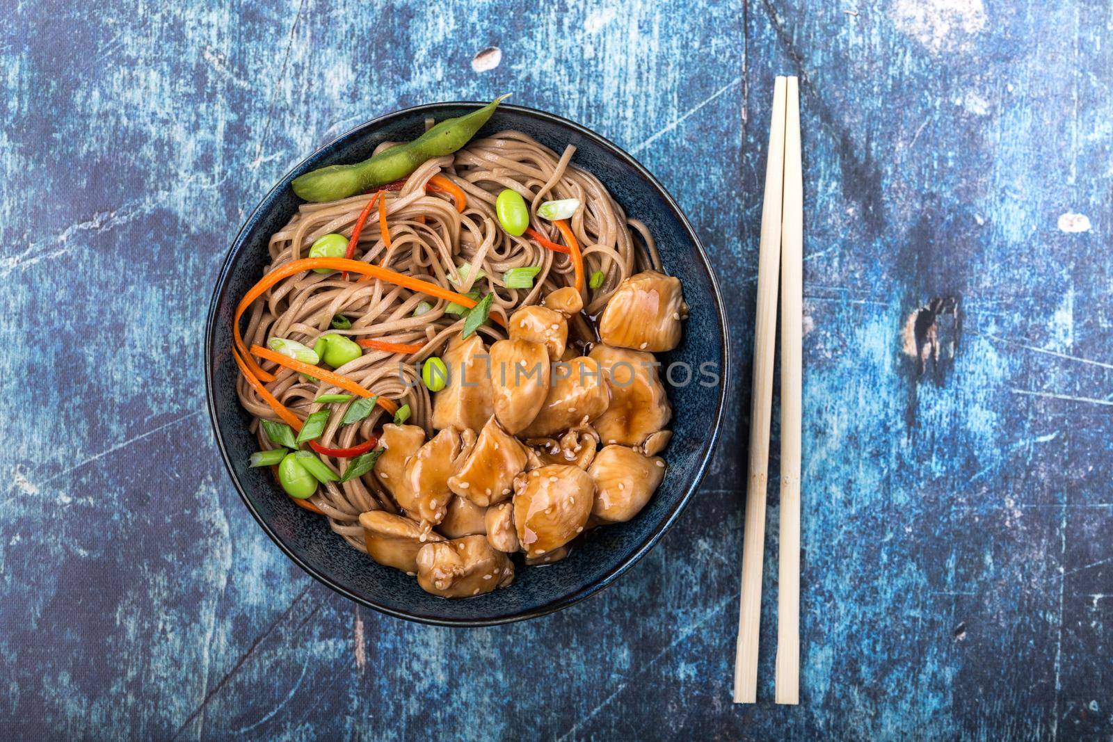 Asian noodles with chicken, vegetables, bowl, rustic wooden blue background. Closeup. Top view. Soba noodles, teriyaki chicken, edamame, chopsticks. Asian style dinner/lunch. Chinese/Japanese noodles