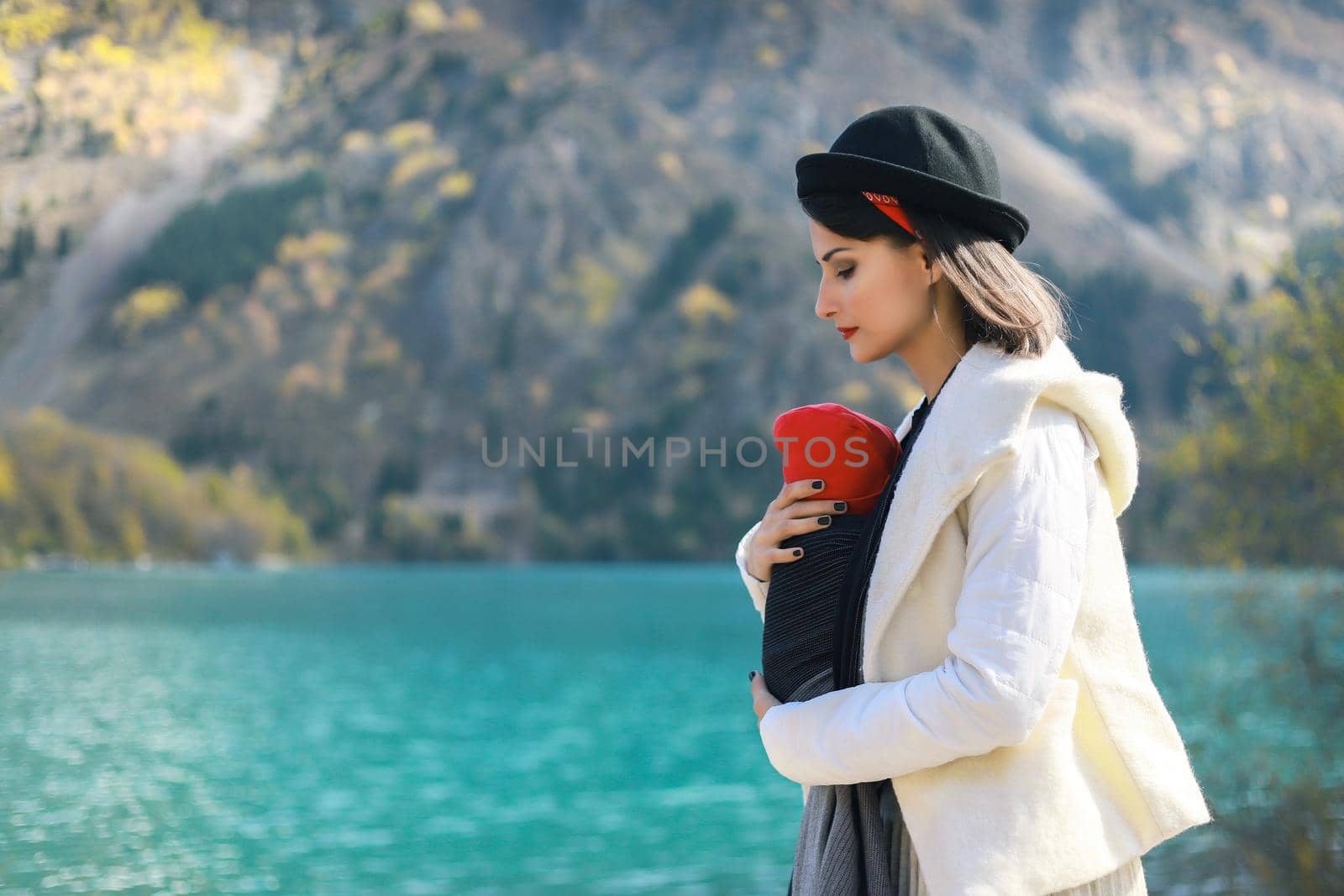 Beautiful young mother with her infant baby in sling outdoor. Mother is carrying her child and travel in autumn mountain lake. Babywearing concept