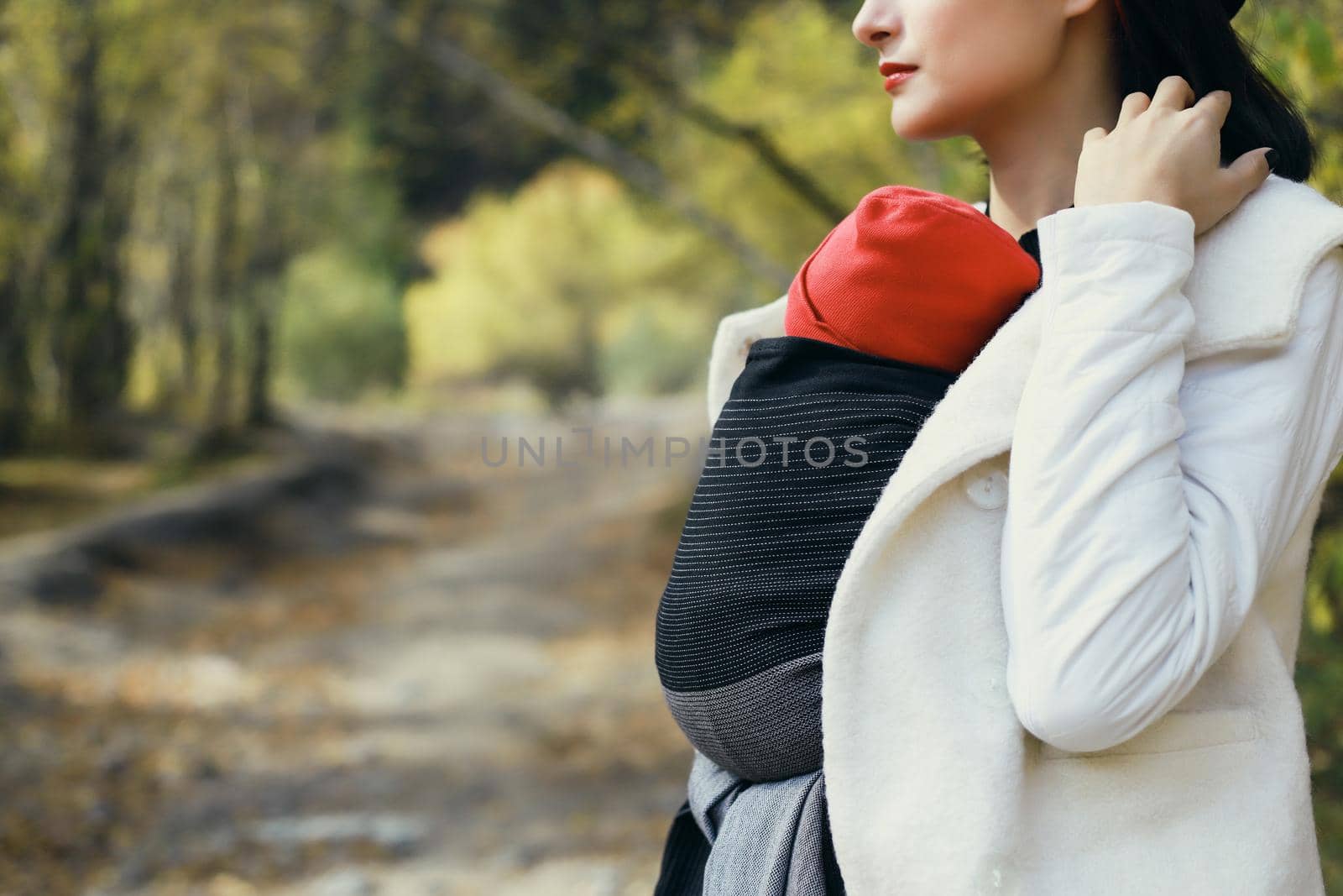 Beautiful young mother with her infant baby in sling outdoor. Woman is carrying her child and walk in autumn nature. Babywearing concept by Rom4ek