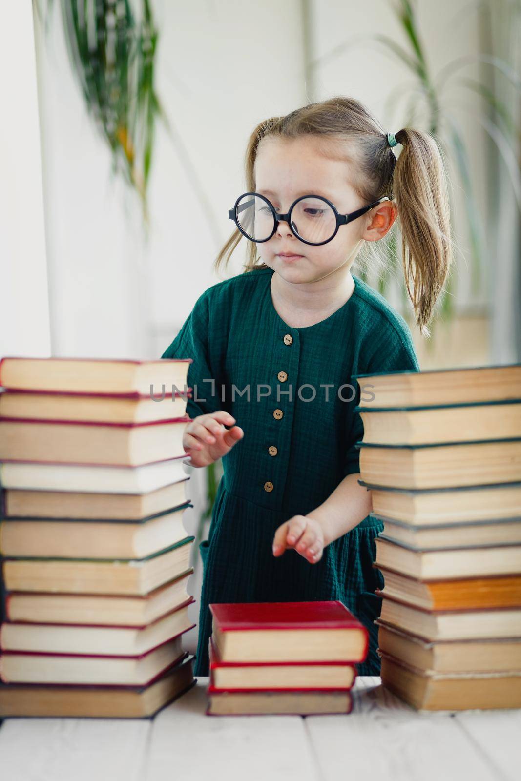 Cute little girl in round shaped glasses and green muslin dress folds colorful books in order.