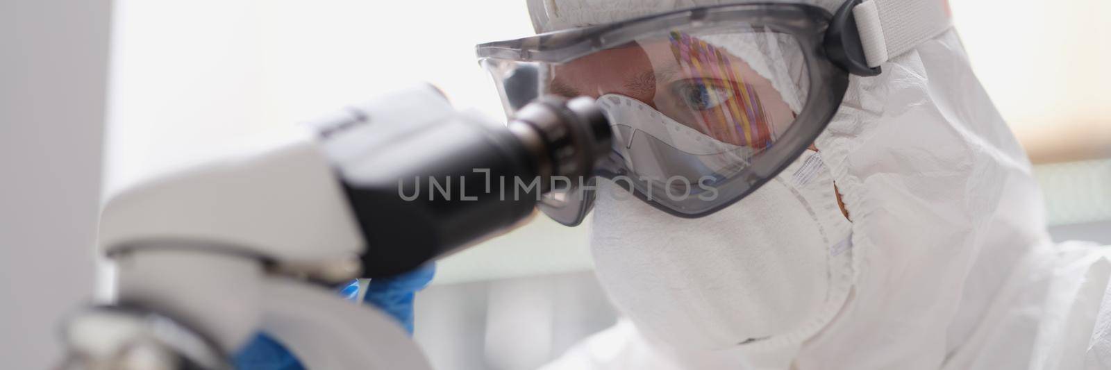 A man in a protective suit and mask looks through a microscope by kuprevich