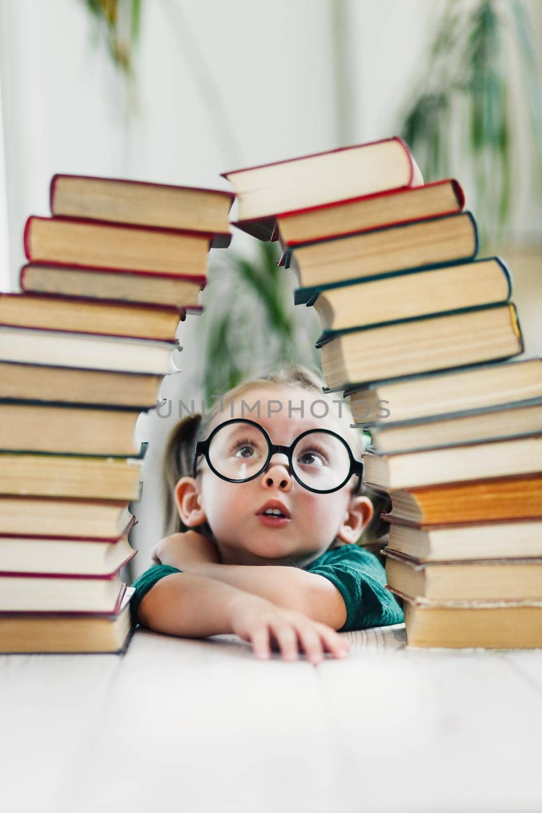 Cute little girl in round shape glasses and green dress under arch made of books. Reading and kids education concept.