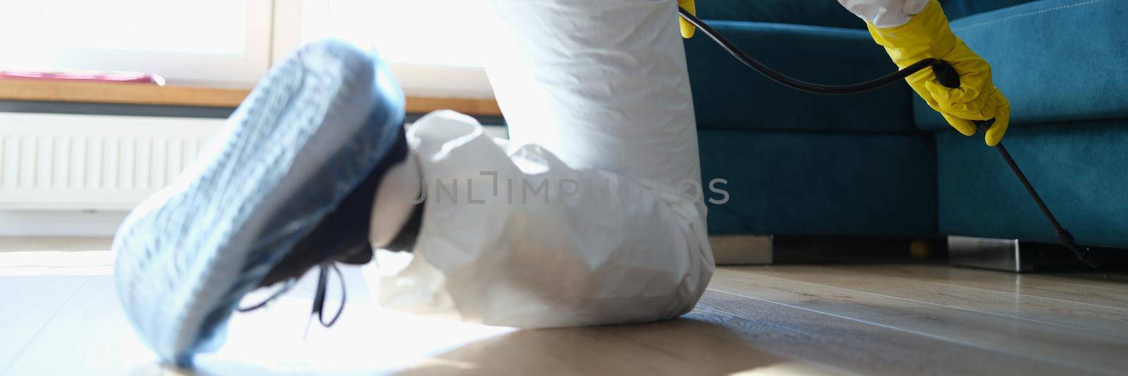 A man in a protective suit sprays under the sofa, close-up. Rodent protection, antiviral chemical treatment