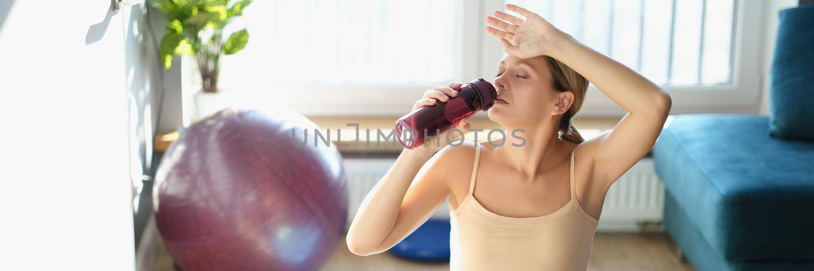 Tired woman drinks water while sitting on the floor with a laptop. Online fit training, sports blog, athletic body