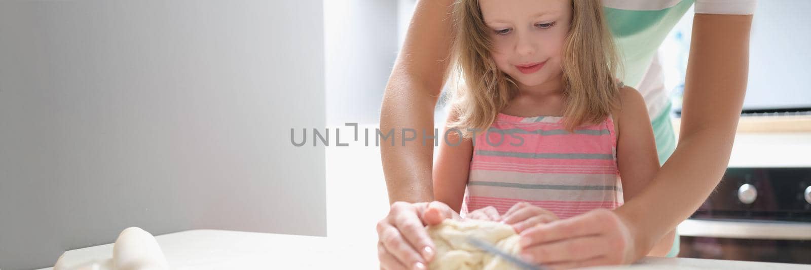 Mom and daughter are kneading dough together in the kitchen, close-up. A little girl learns from her parents how to cook food at home