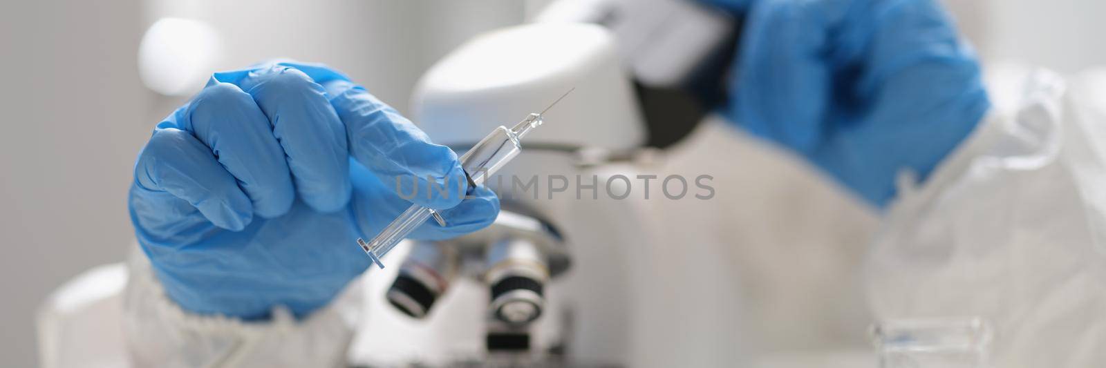 A man holds a syringe with a liquid and looks through a microscope, close-up, blurry. Virus drug research laboratory