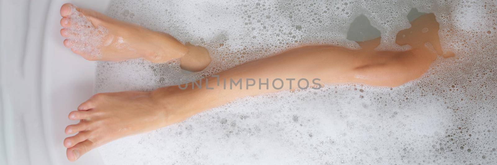 Slender female legs in a bathtub with soapy water, close-up. Bath foam, home aromatherapy, bath salt