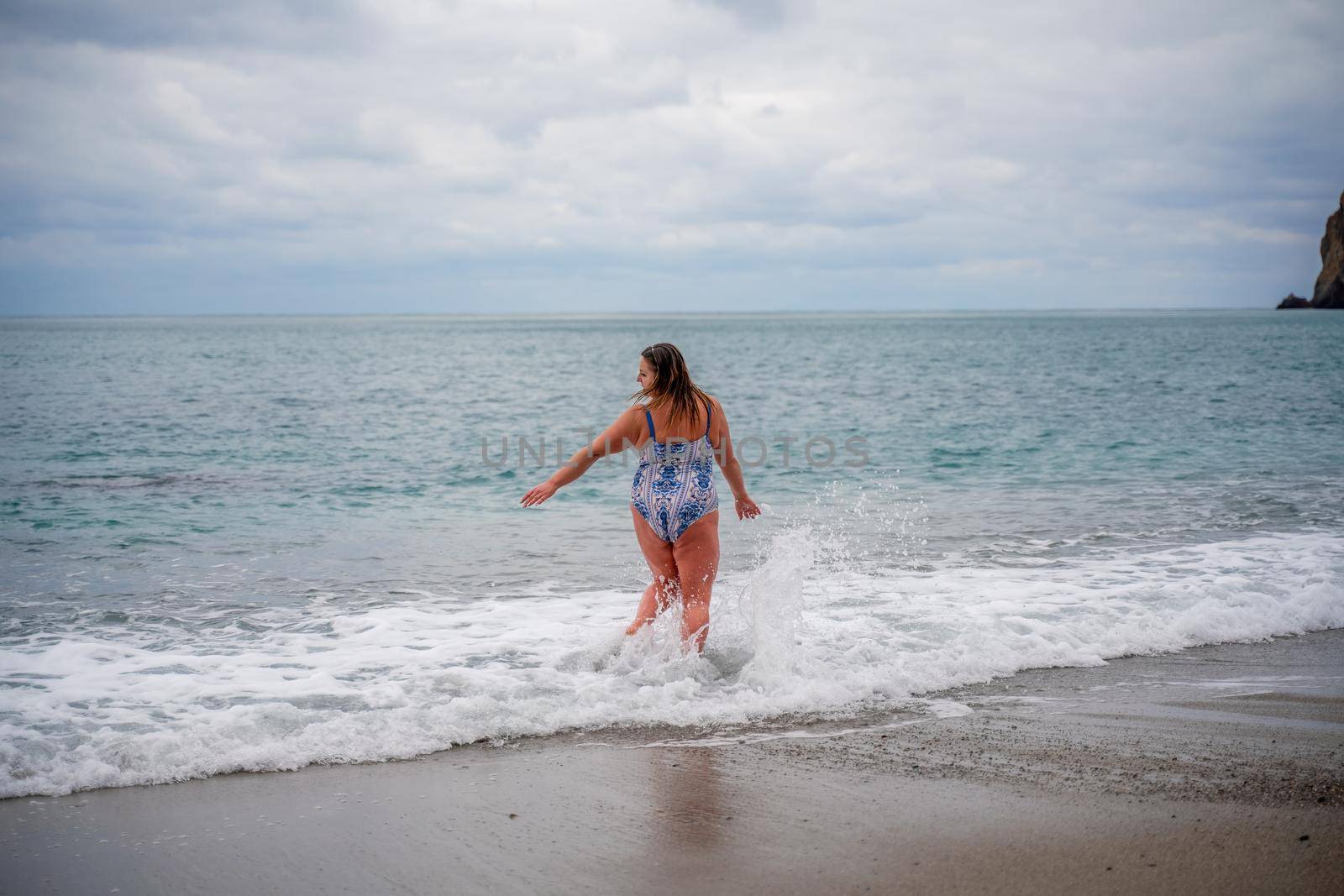 A plump woman in a bathing suit enters the water during the surf. Alone on the beach, Gray sky in the clouds, swimming in winter