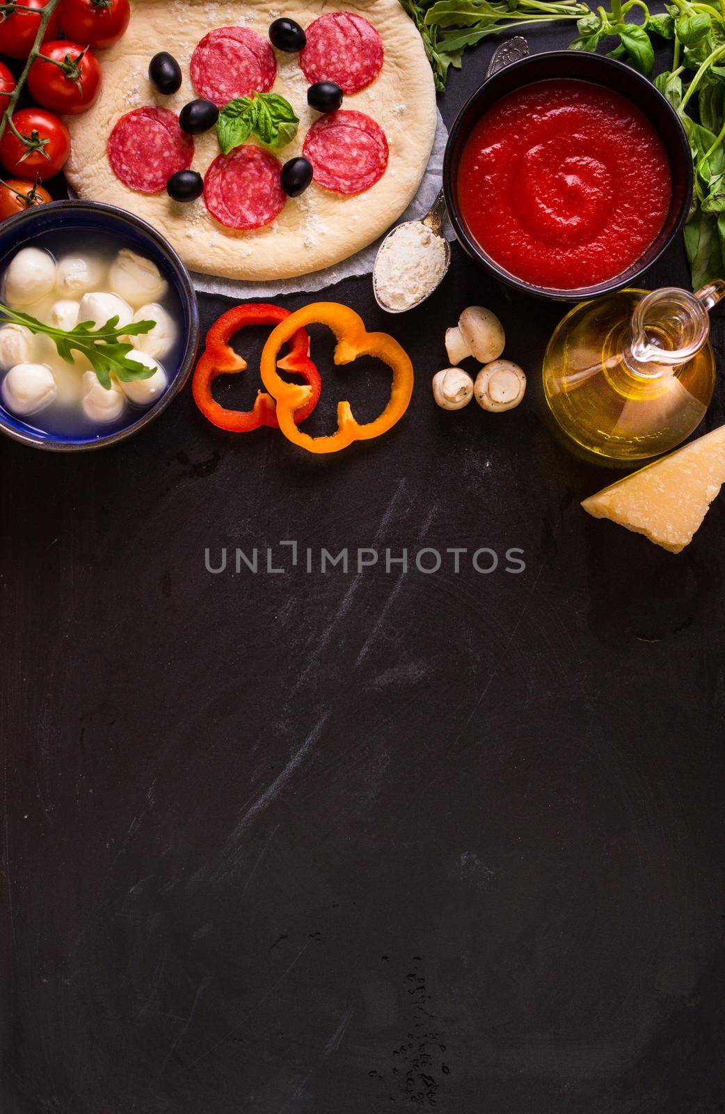 Pizza making background. Ingredients for making pizza. Space for text. Pizza dough, flour, cheese, mozzarella, tomatoes, basil, pepperoni, olives and rolling pin over black chalkboard. Top view