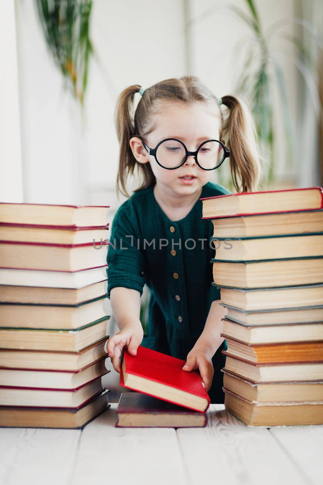 Cute little girl in round shaped glasses and green muslin dress folds colorful books in order.