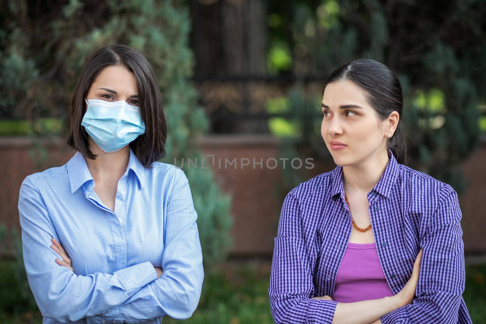 Two young women sits on a summer park bench. One woman wearing a mask, one without a mask. Different attitudes to pandemic.